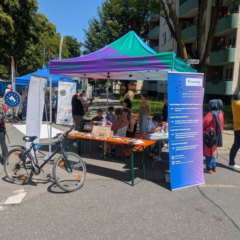 Marktstand mit buntem Zelt, Tischen und Fahrrädern, umgeben von Menschen.